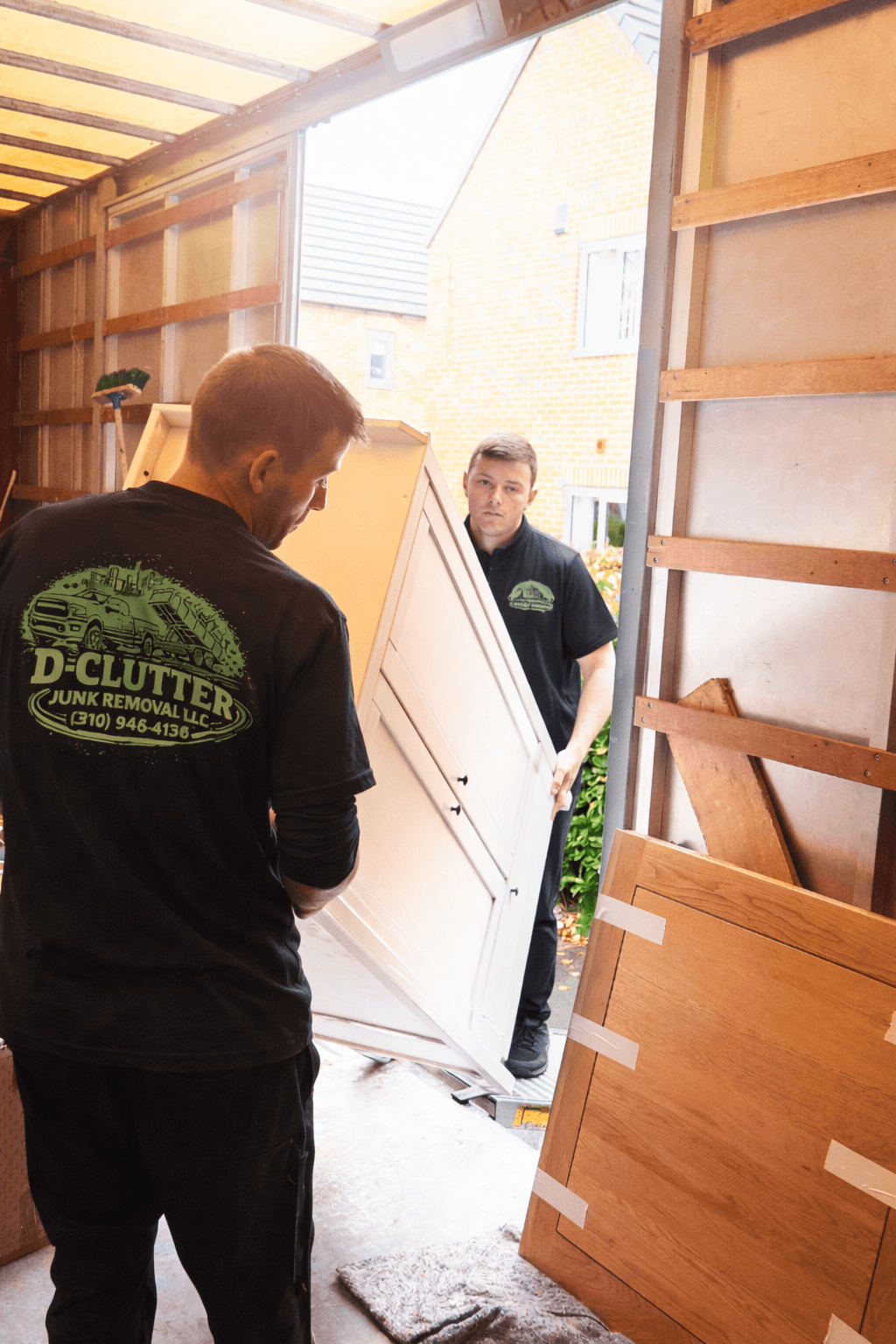 Two men from D-CLUTTER Junk Removal loading a white cabinet into a truck.