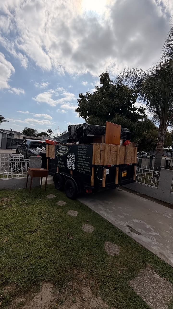 Black junk removal trailer filled with items parked on a driveway beside a small table.