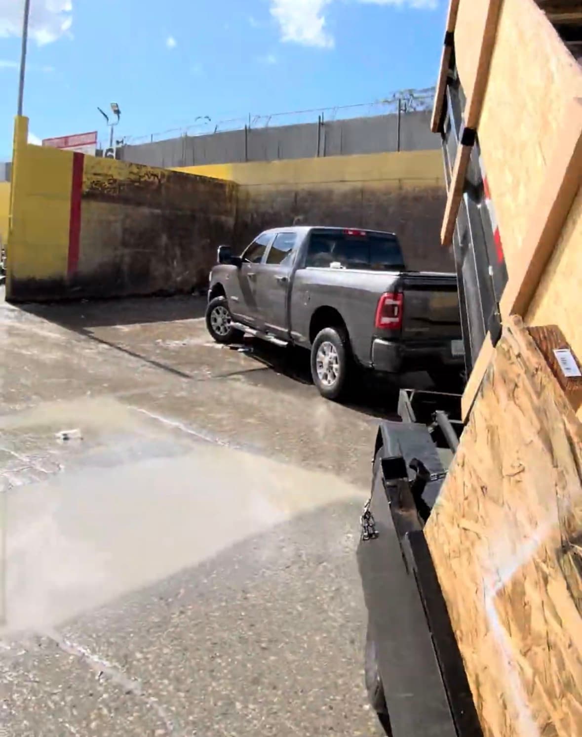 Grey pickup truck parked in an industrial lot beside a yellow wall and large puddle.