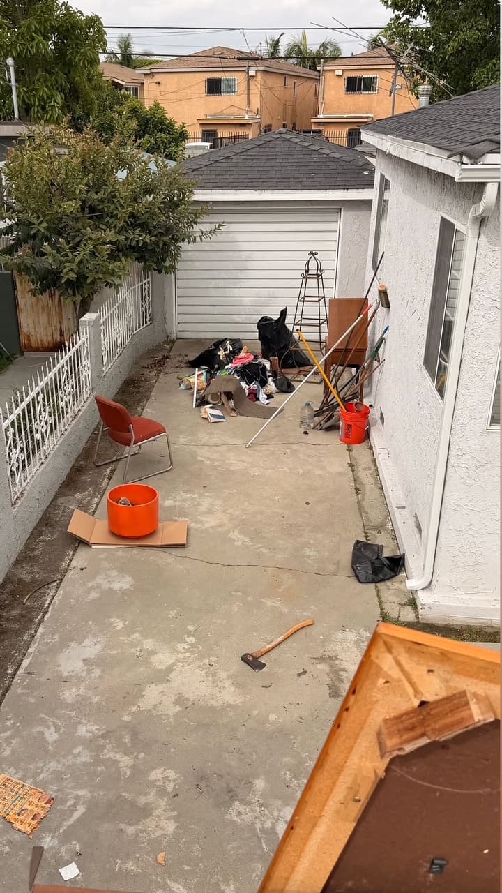 Narrow concrete driveway cluttered with an axe, red chair, orange bucket, and various debris.