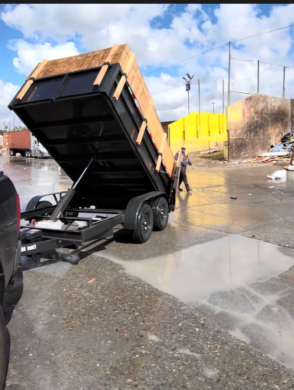 Black hydraulic dump trailer with wooden sideboards tilted up at a wet industrial site.